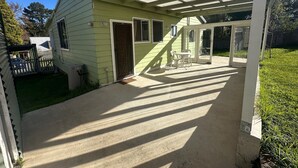 Ein überdachter Patio-Bereich mit Betonboden, einem kleinen Tisch mit Stühlen und einer Glasschiebetür, die in einen Garten führt.