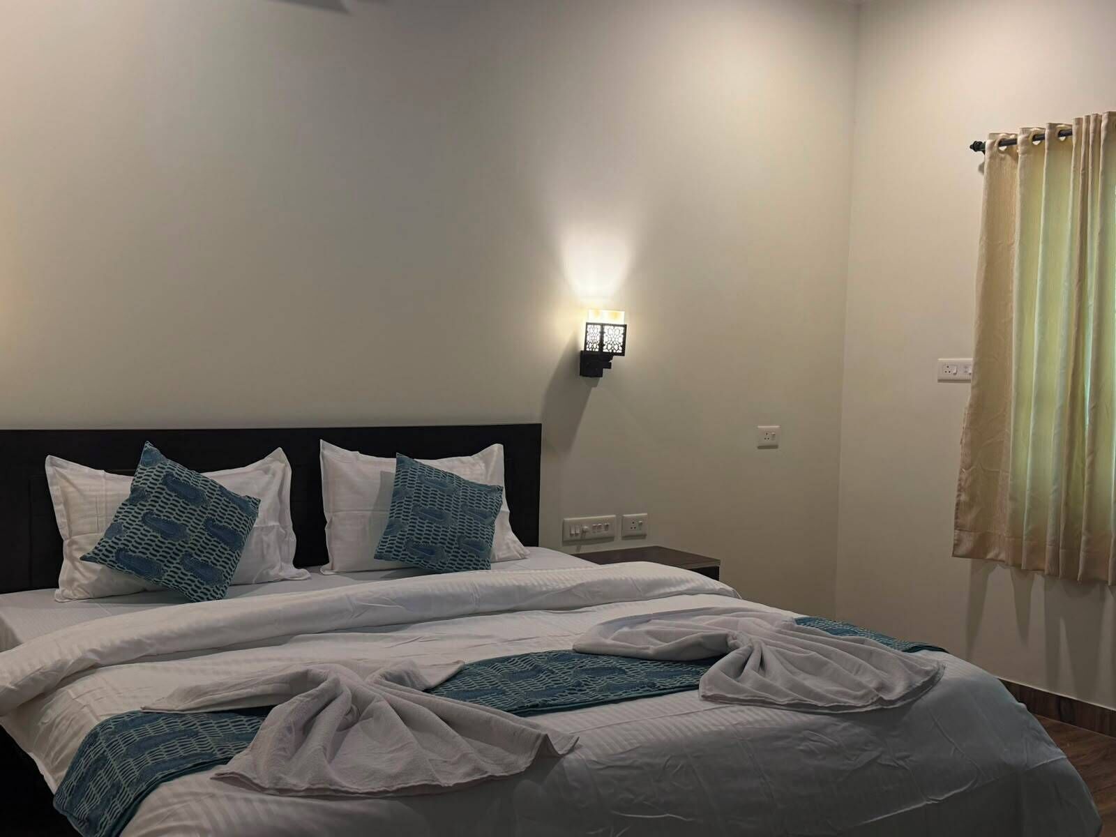 Una habitación de hotel con cama, ropa de cama blanca, una lámpara de pared y una ventana con cortinas.