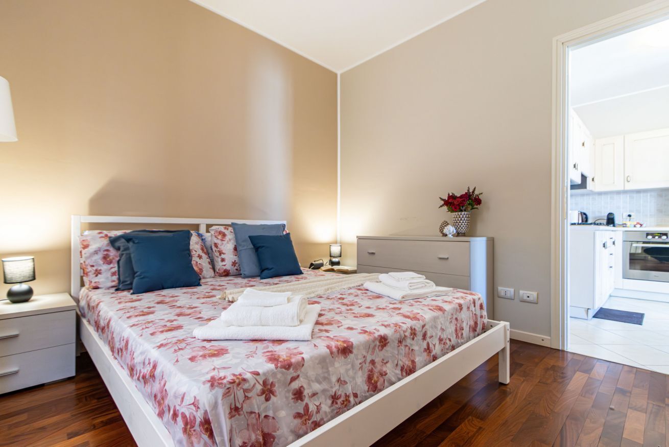 A bedroom with a bed, bedside tables, a nightstand, a dresser, and a view of the kitchen.