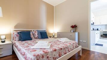 A bedroom with a bed, bedside tables, a nightstand, a dresser, and a view of the kitchen.