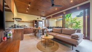 A modern kitchen with wooden cabinets, a dining area with a table and chairs, a sofa, and a ceiling fan. - Nala Villa Bali (Badung)