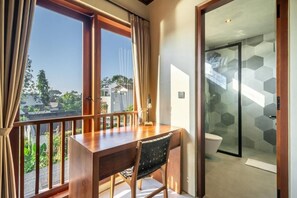 A room with a wooden desk, a chair, a view of greenery outside, and a glass shower with hexagonal tiles. - Nala Villa Bali (Badung)