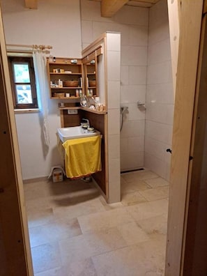 A small, compact bathroom with a shower, a sink with a yellow towel, and a wooden shelf unit with toiletries. - Hüthis Waldidylle (Edlbach)