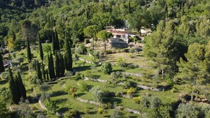 Une propriété rurale dotée de jardins en terrasses, d’une maison en pierre avec un toit en tuiles, et entourée d’une forêt dense.