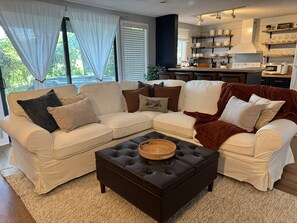 Ein Wohnzimmer mit Ecksofa, dunklem Hocker und einer Küche im Hintergrund.