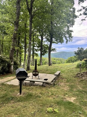 Ein Picknickplatz mit Grill und Sitzgelegenheit, umgeben von Bäumen und Blick auf ferne Hügel.