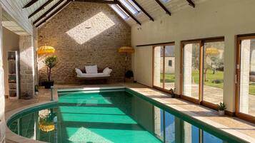 A swimming pool with a stone wall and a wooden ceiling, large windows, and a sunlit interior.
