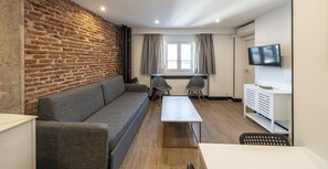 Un salon moderne avec un mur en briques, un canapé gris, une table basse blanche, deux fauteuils, un téléviseur et une fenêtre avec des rideaux.