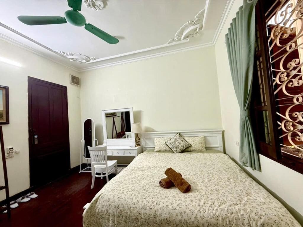 A bedroom with a bed, a desk, a chair, a ceiling fan, and a window with curtains.