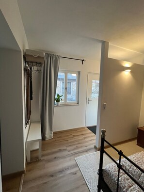 A room with a wooden floor, a bed with a white and floral bedspread, a window with a curtain, and a plant on the windowsill.