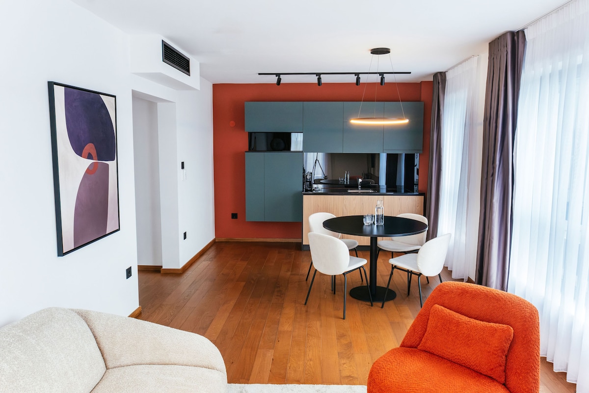 A modern living room with a sofa, a dining table with chairs, a kitchen area with cabinets, and a large window with curtains.