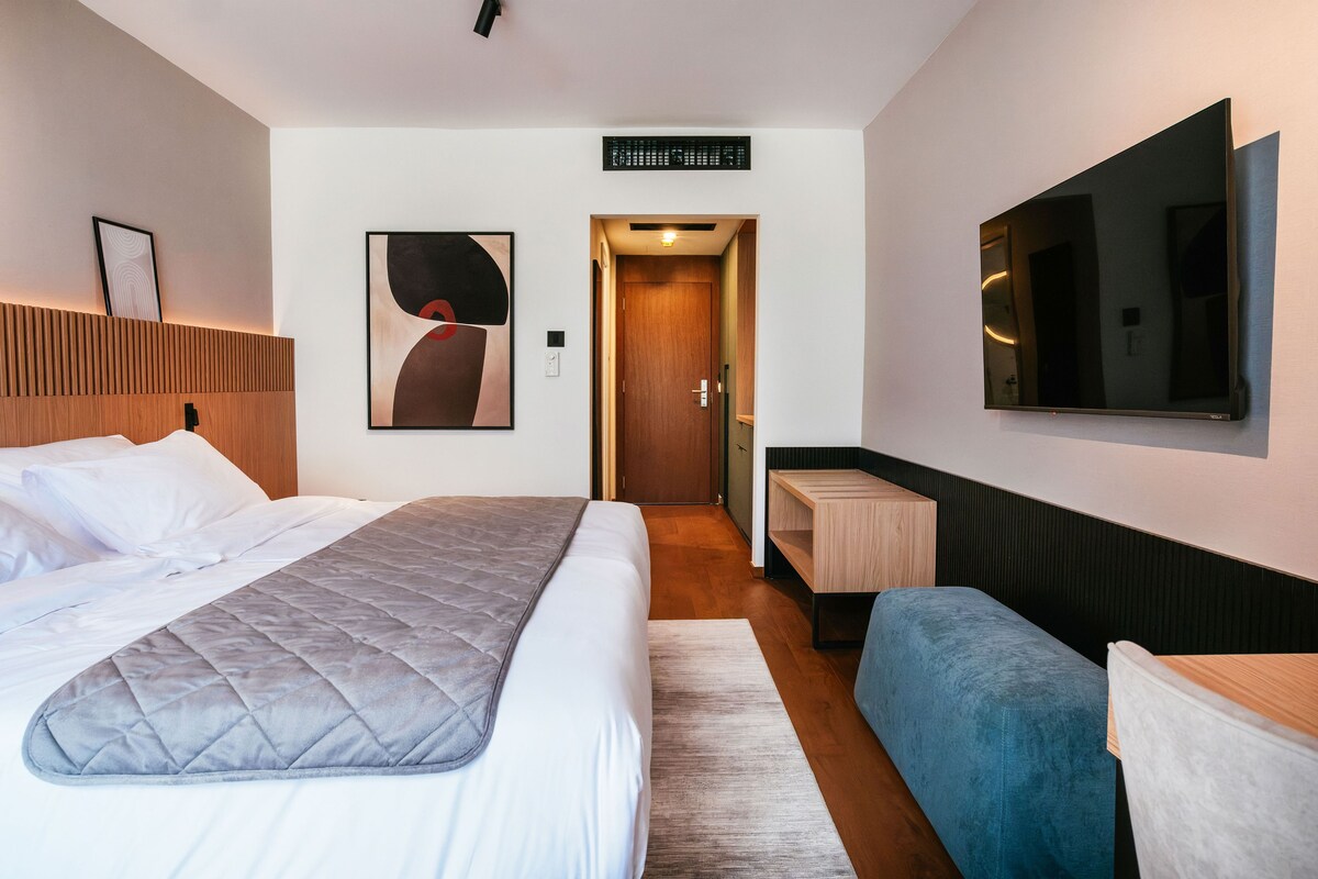 A hotel room with a bed, a bedside table, a seating area, a TV, and a door leading to another room.