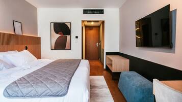 A hotel room with a bed, a bedside table, a seating area, a TV, and a door leading to another room.