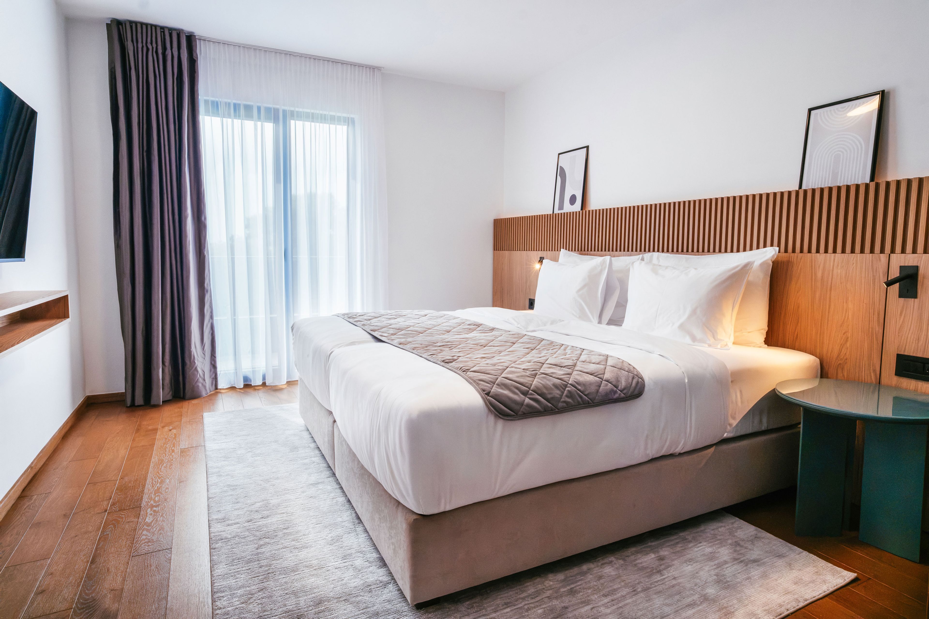 Une chambre d’hôtel moderne avec un grand lit, une tête de lit en bois, une table de chevet et une vue sur la ville par la fenêtre.