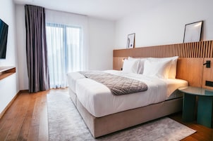 Una habitación de hotel moderna con una cama grande, cabecera de madera, mesita de noche y vistas a la ciudad por la ventana.