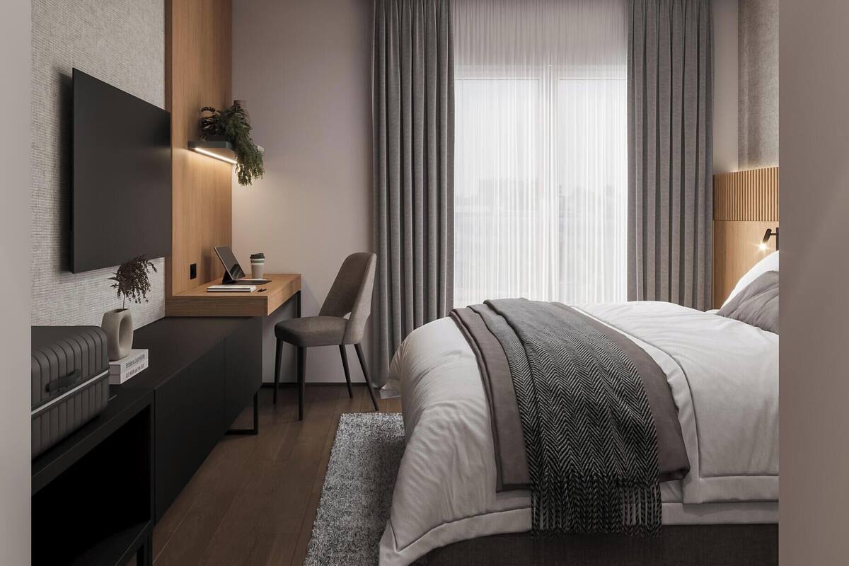 Moderni hotellihuone, jossa on taulutelevisio, työpöytä kannettavalla tietokoneella, mukava tuoli, suuri sänky ja ikkuna, jossa on verhot.
