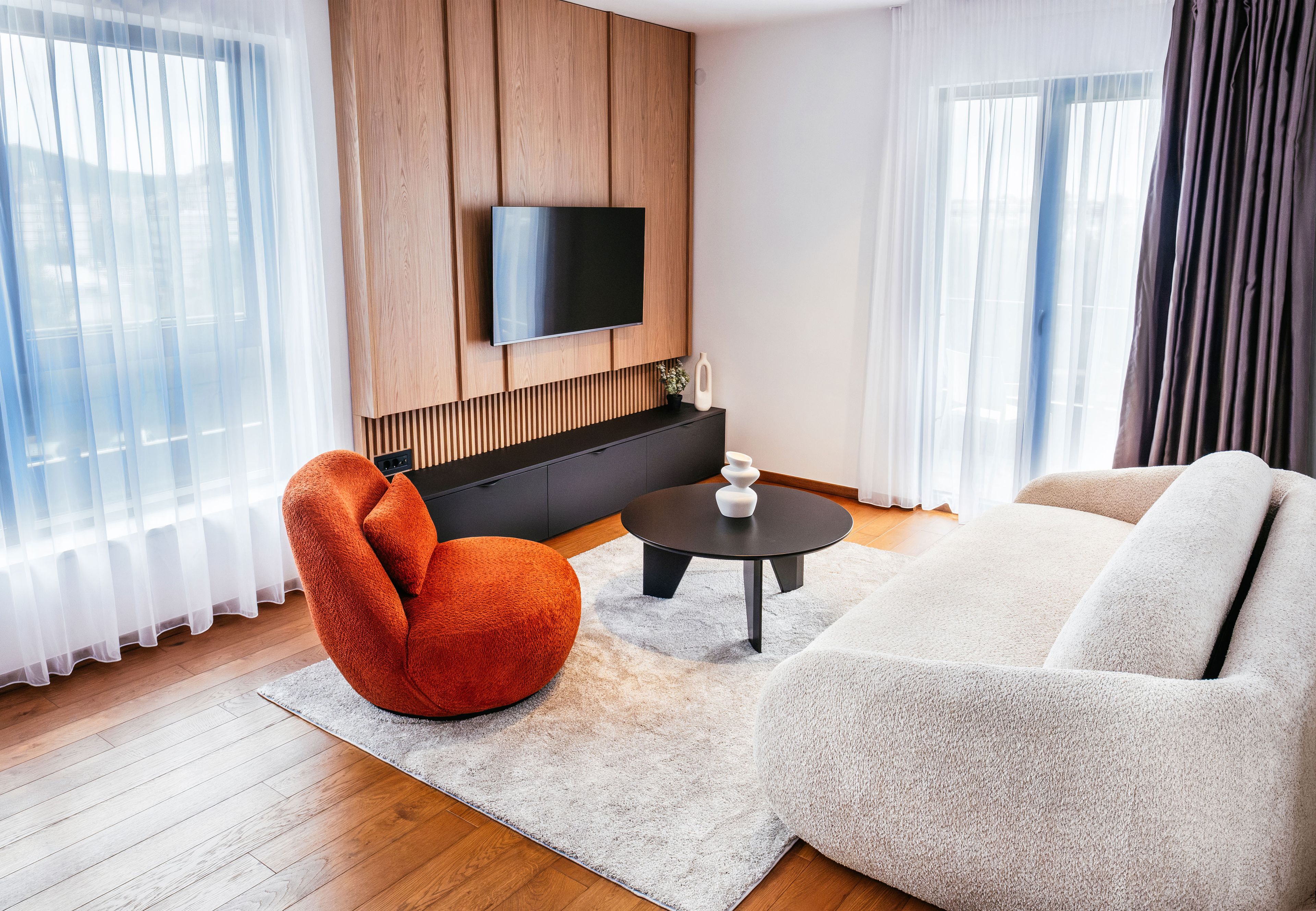 Una sala moderna con un televisor de pantalla plana montado en una pared de madera, un sillón rojo, un sofá blanco y una pequeña mesa redonda.