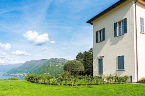 Ein Gebäude mit weißer Fassade und blauen Fensterläden, umgeben von einer gepflegten Rasenfläche und Blick auf einen See und Berge.