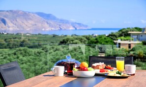 A table with breakfast items and a glass of orange juice, overlooking a scenic landscape with mountains and the sea. - Minoas Sky Villa Heated Pool (Lake Kournas)