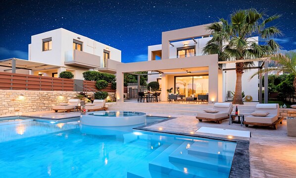 A modern outdoor pool area with a clear night sky, a stone wall, and lounge chairs. - Minoas Sky Villa Heated Pool (Lake Kournas)