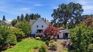 Uma casa de dois andares com gramado bem cuidado, um alpendre com móveis de exterior e uma grande árvore no jardim da frente.