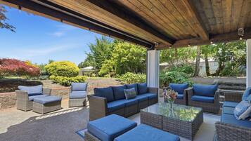 An outdoor patio with wicker furniture and a glass table, surrounded by a garden and trees.