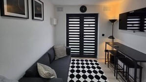 Ein moderner Wohnraum mit Sofa, einem gemusterten Teppich, einem Fernseher und einer Tür mit horizontalen Jalousien.