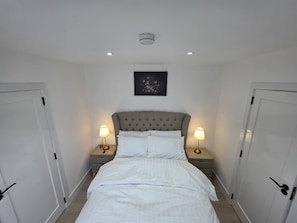 A bedroom with a tufted headboard, white bedding, two bedside lamps, a framed artwork above the bed, and white doors on either side.