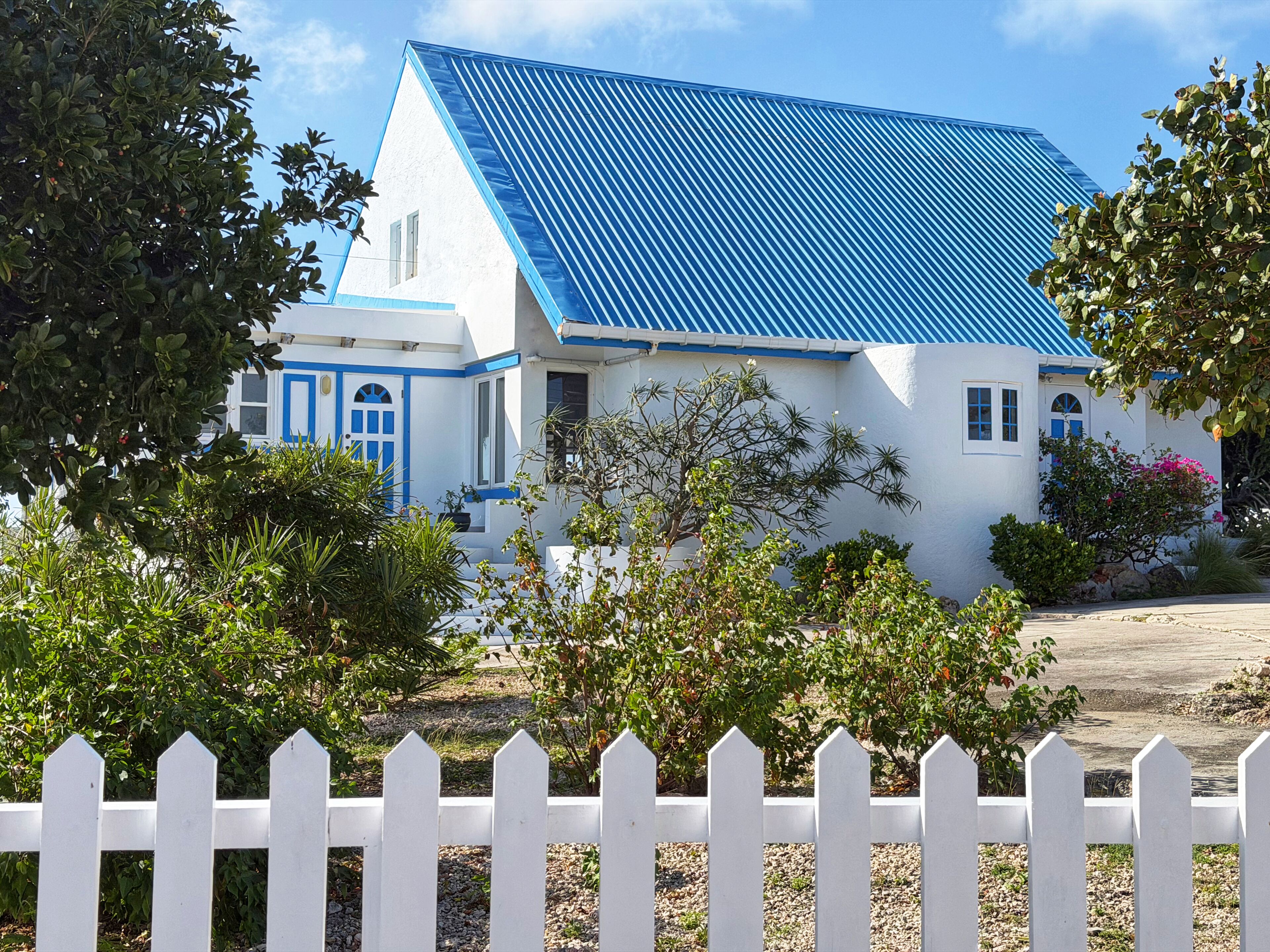 Un bâtiment blanc surmonté d’un toit bleu, entouré de verdure et d’une clôture blanche en bois.