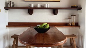 Ein Essbereich mit einem Holztisch, Hockern, einer Bananenschale, einem Bild darüber und Regalen mit Gegenständen.