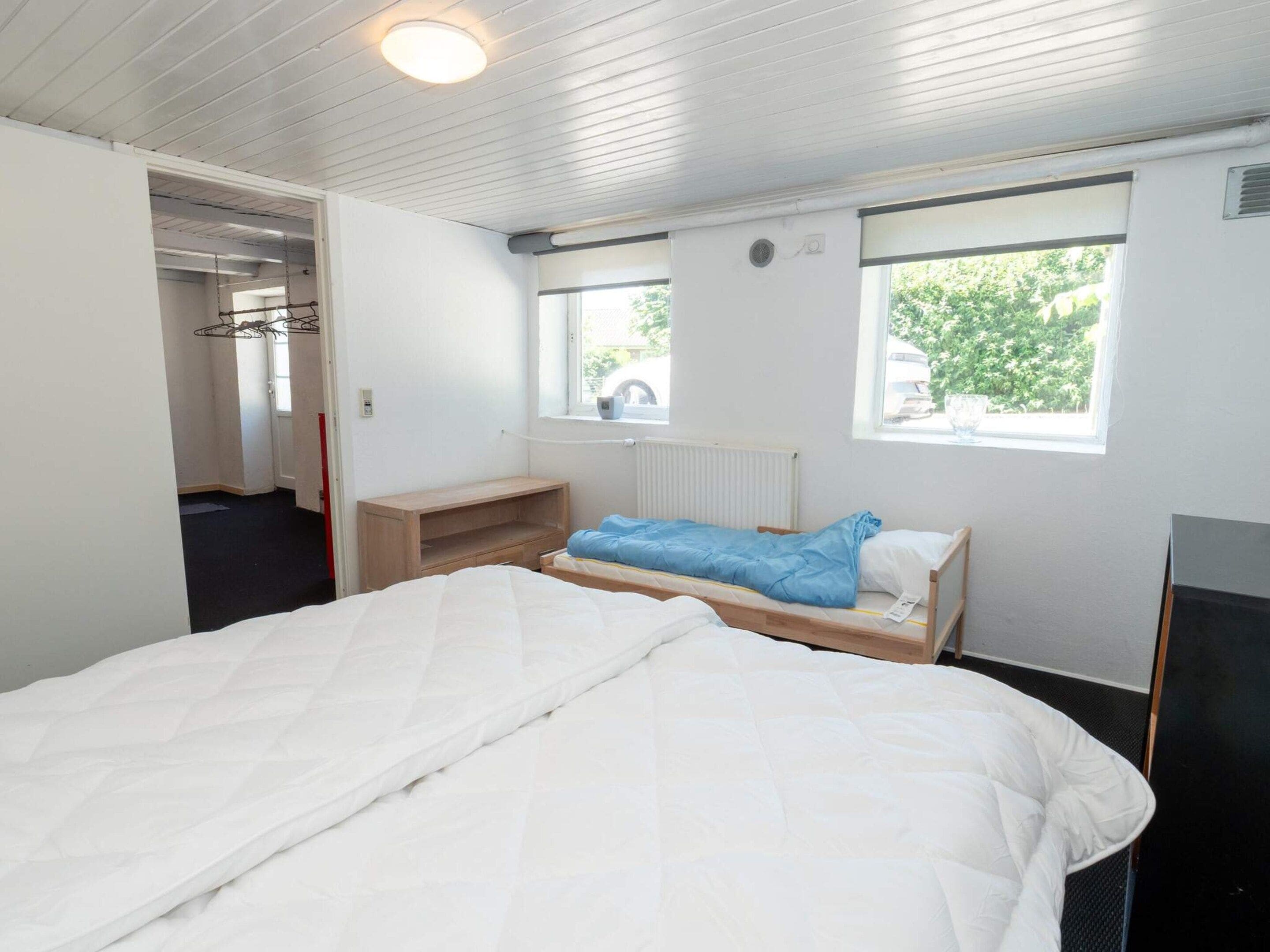 Una cama individual con edredón blanco, una mesita de noche de madera, una manta azul y una ventana con vistas a la vegetación.