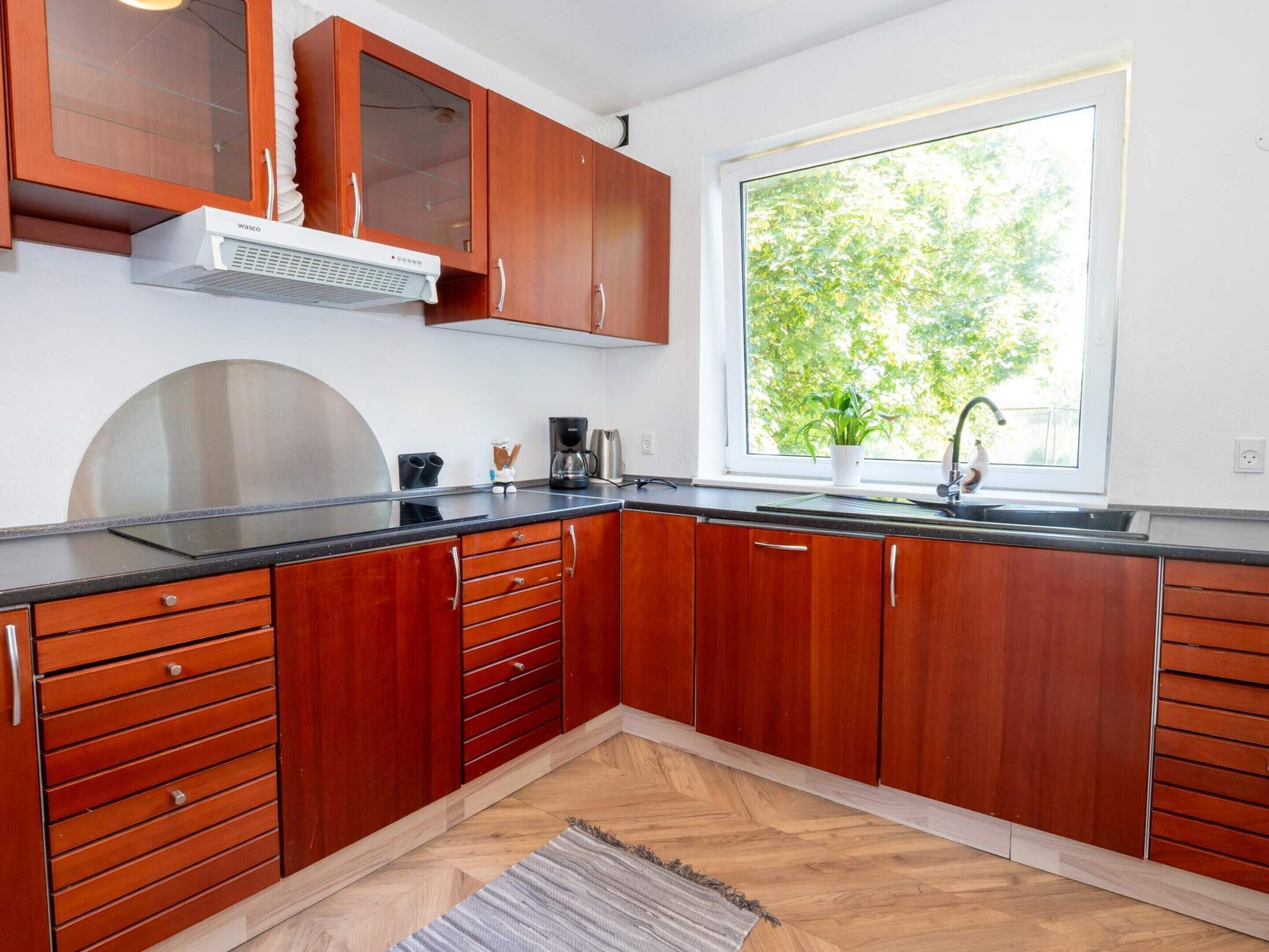Una cocina moderna con armarios de madera, extractor de acero inoxidable, encimera negra y ventana con vista a árboles.