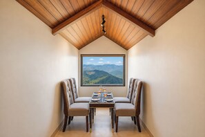 A dining area with a table set for two, a large window with a mountain view, and wooden ceiling beams.