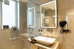 A modern bathroom with a glass shower enclosure, a white sink, a mirror, and a hairdryer on the wall. - ELIVAAS Privé Royce Silver Oak APT (Kasauli)