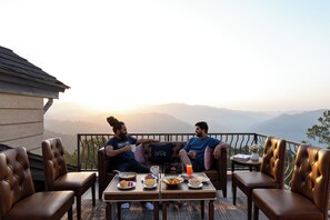 Two men sitting on a balcony with a table of breakfast items, a view of mountains, and a cozy seating area. - ELIVAAS Privé Royce Silver Oak APT (Kasauli)