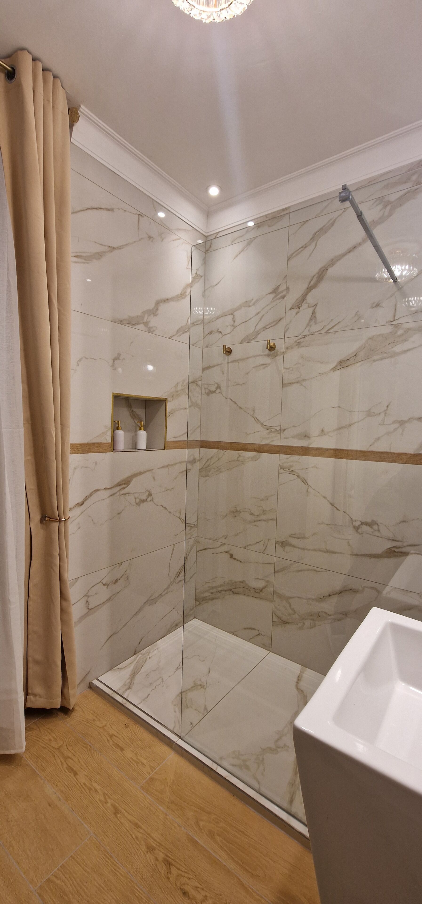 Ein modernes Badezimmer mit Duschkabine aus Glas, Marmorwänden, Holzboden und Waschbecken in Weiß.