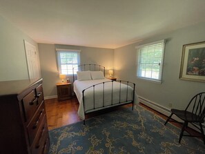 Ein Schlafzimmer mit Bett, Nachttisch, Kommode, Stuhl und zwei Fenstern mit Jalousien.