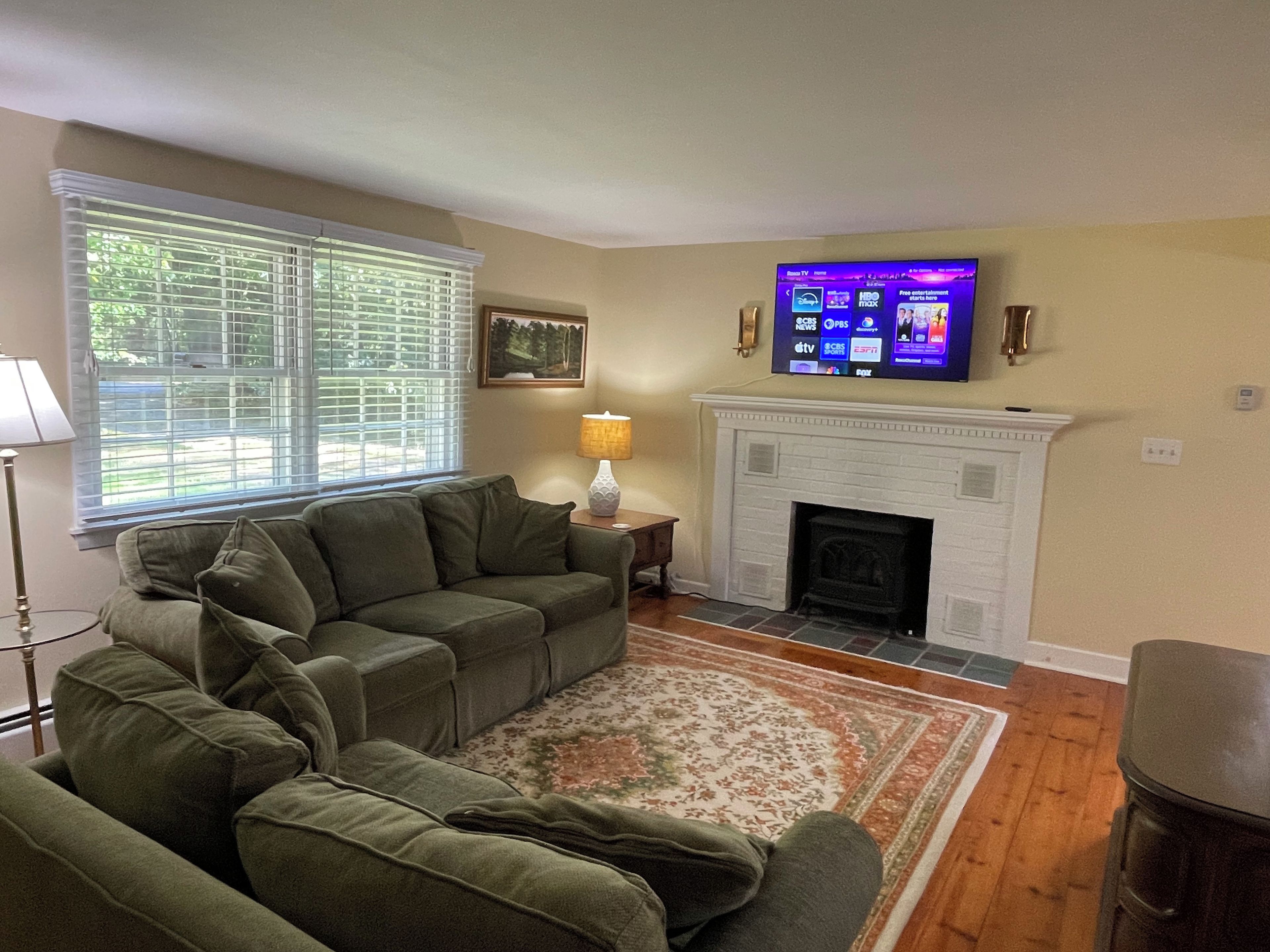 A living room with a sectional sofa, a fireplace, a flat-screen TV mounted above the fireplace, and a window with blinds.