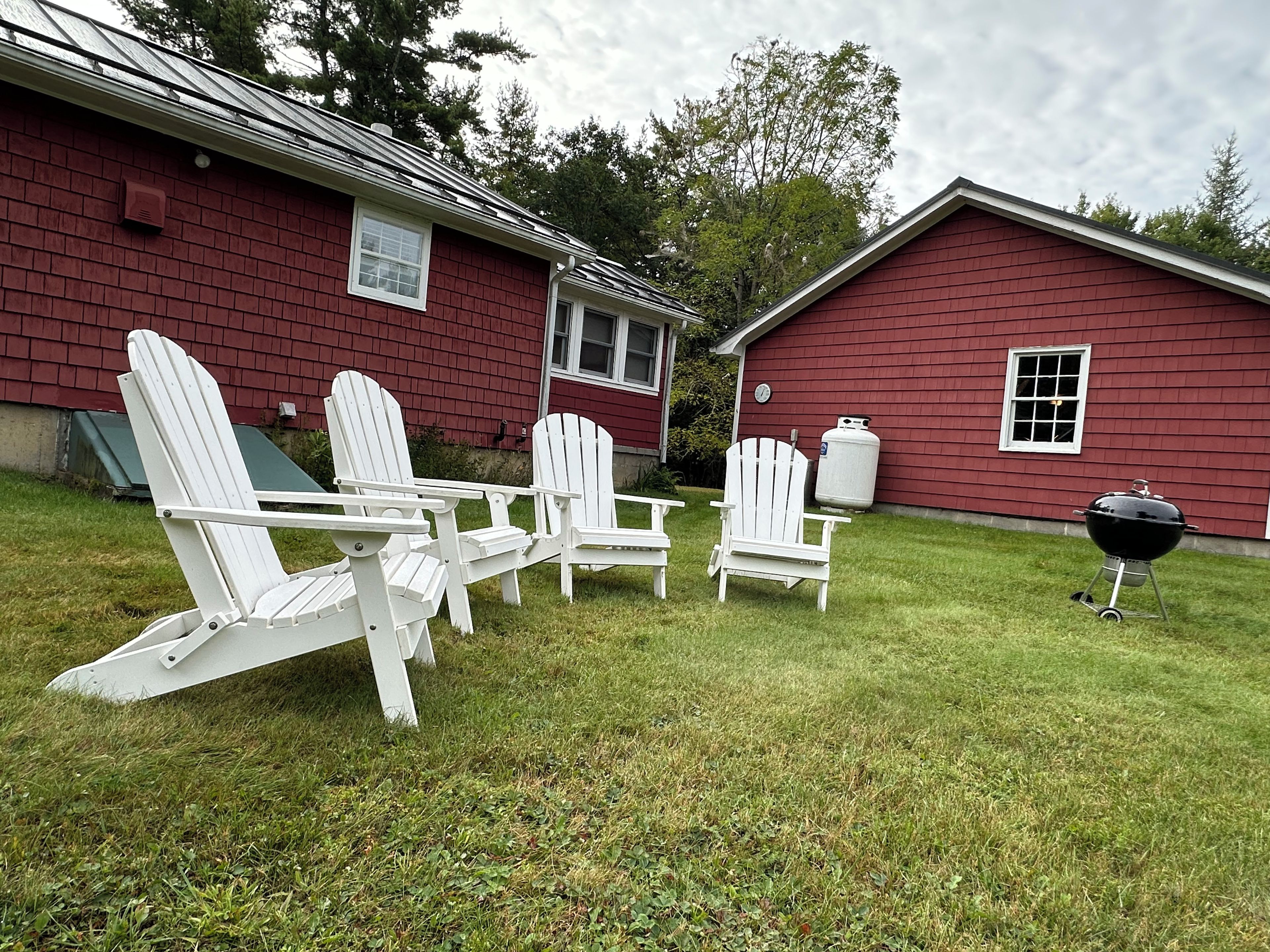 A red building with a black grill and white Adirondack chairs in the yard.