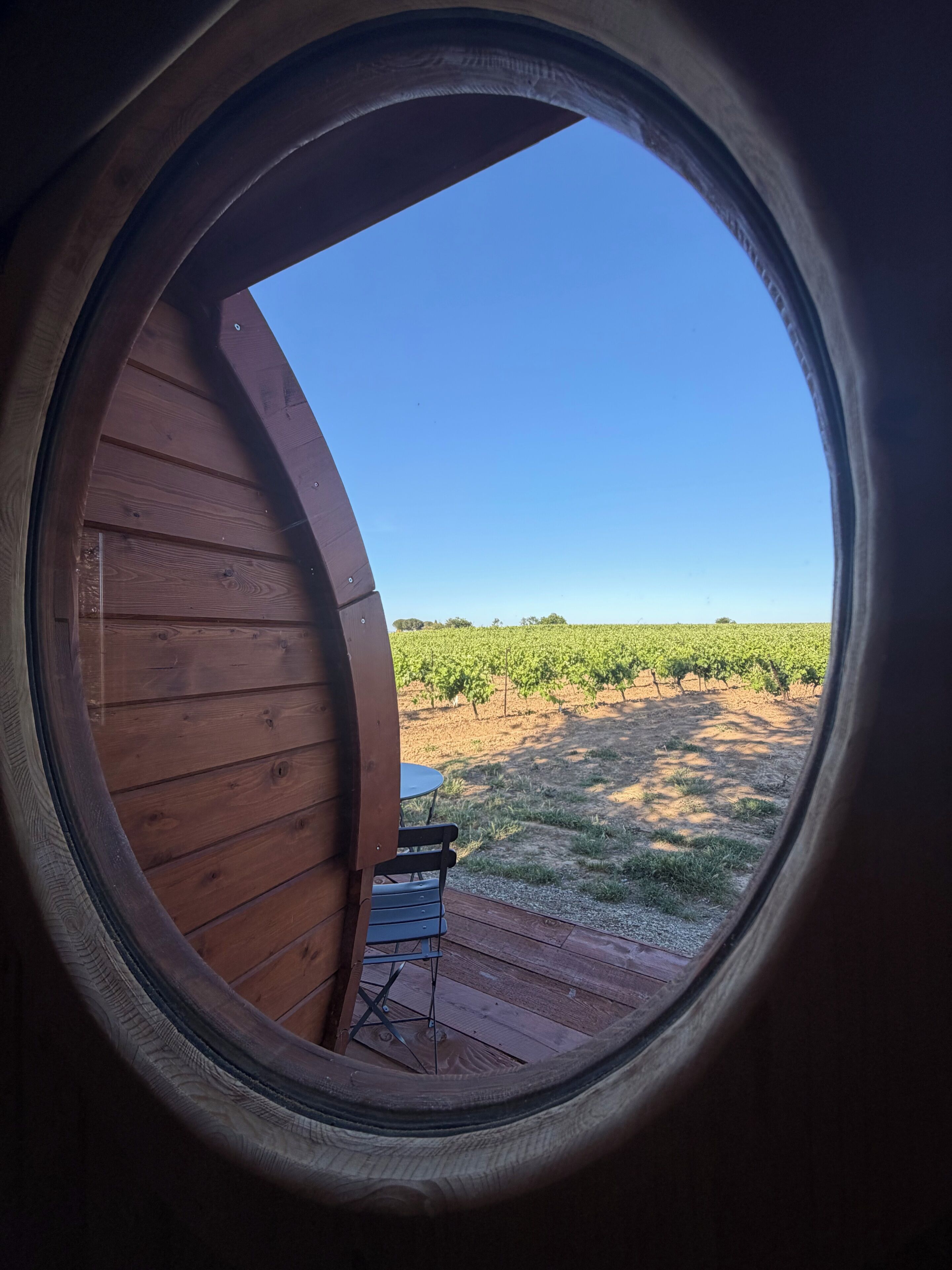Une fenêtre circulaire donnant sur un vignoble et une petite table avec des chaises à l’intérieur.