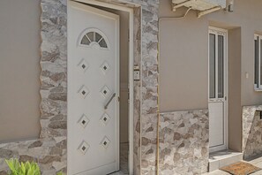 Eine weiße Tür mit rundem Fenster und rautenförmigen Aussparungen, eingelassen in eine Steinmauer mit einem Steinstufenaufgang.
