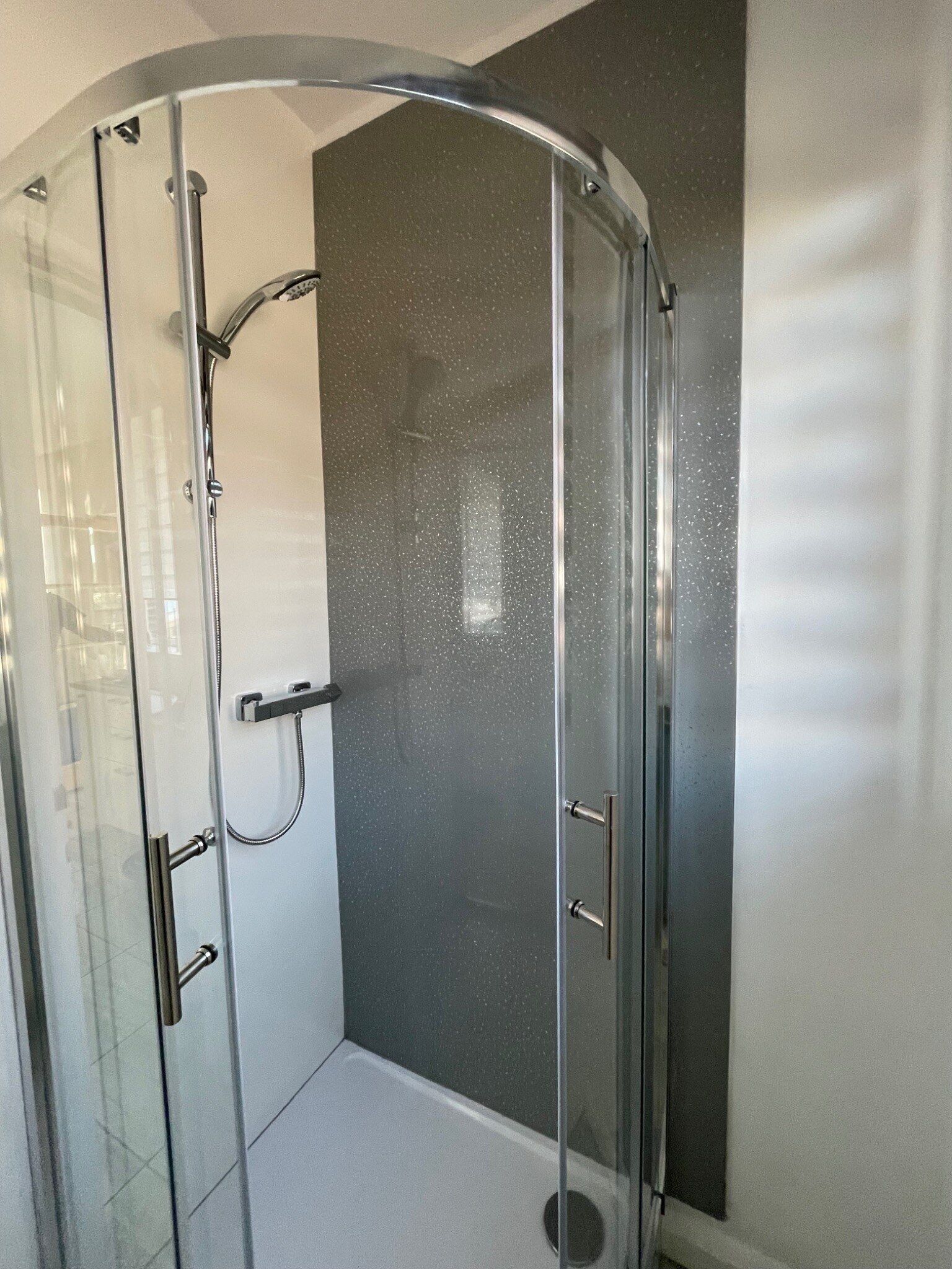 Une douche fermée par des parois en verre, dotée d’une porte incurvée, d’un pommeau de douche à main et d’un panneau de commande fixé au mur.