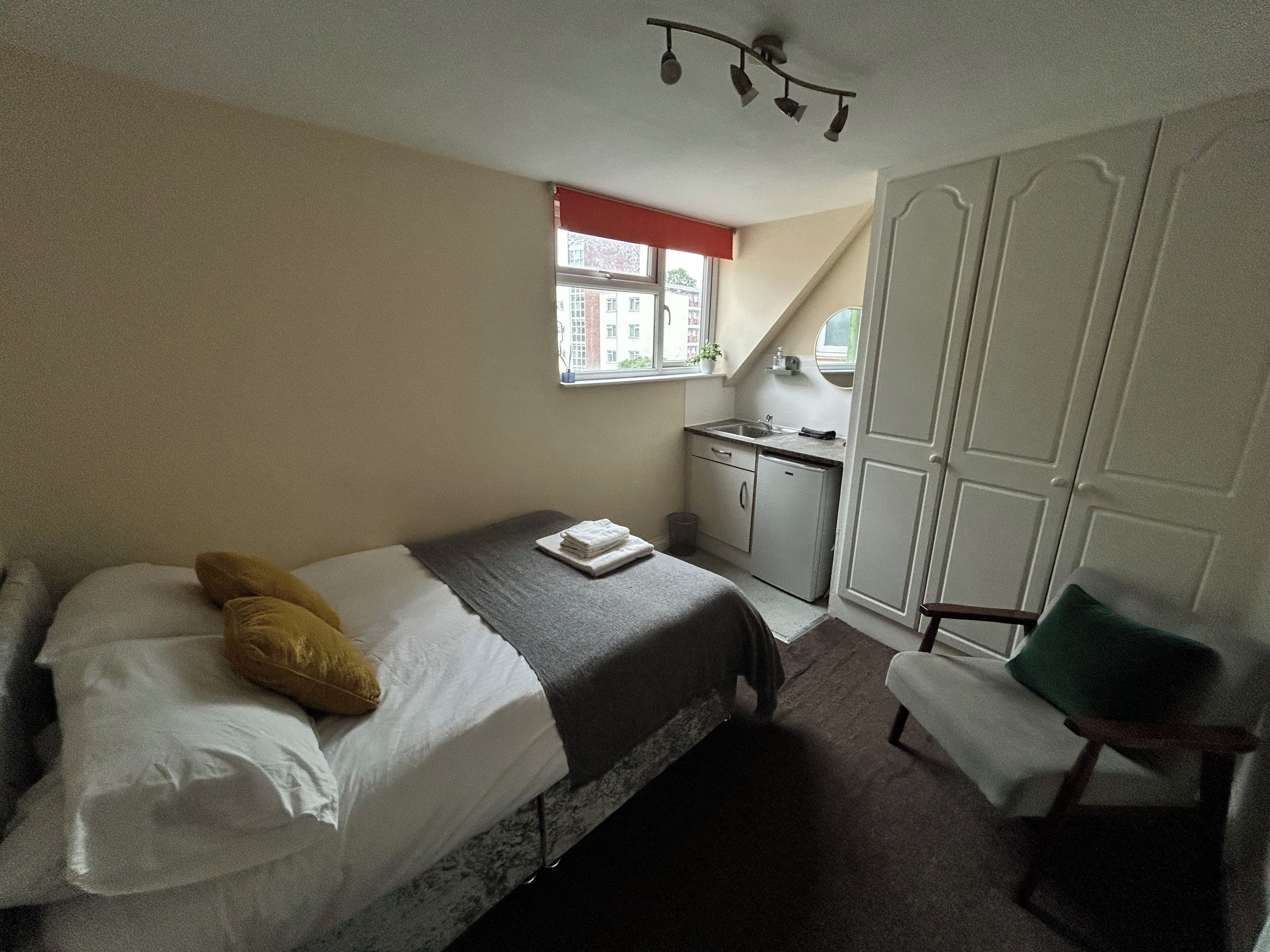A bedroom with a bed, a small kitchenette, a window, and a wardrobe.