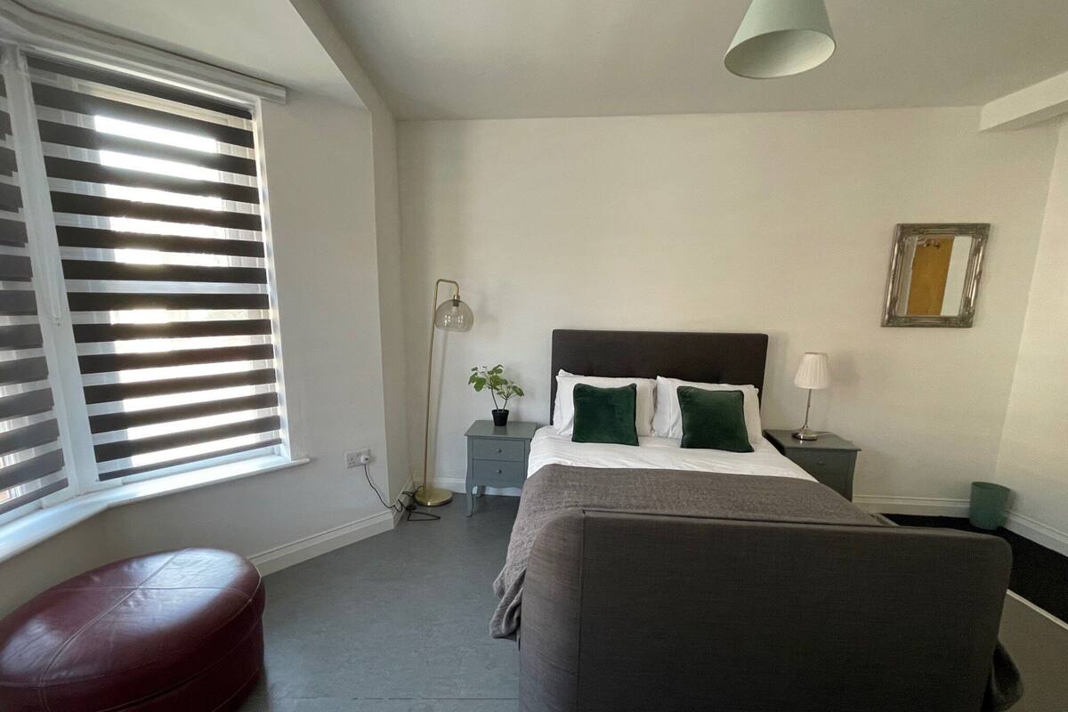 Un dormitorio con cama, mesita de noche, lámpara, planta en maceta y un taburete rojo.