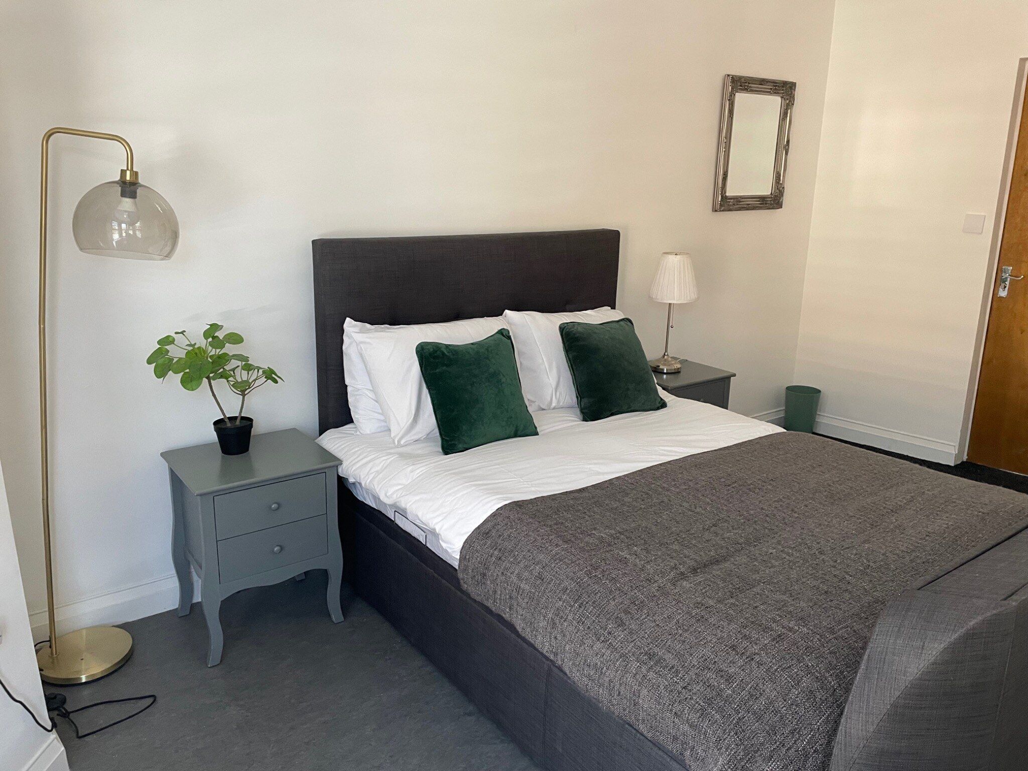 Un lit bien fait, avec une tête de lit sombre, une lampe de chevet, une petite table sur laquelle est posée une plante, et un miroir accroché au mur.