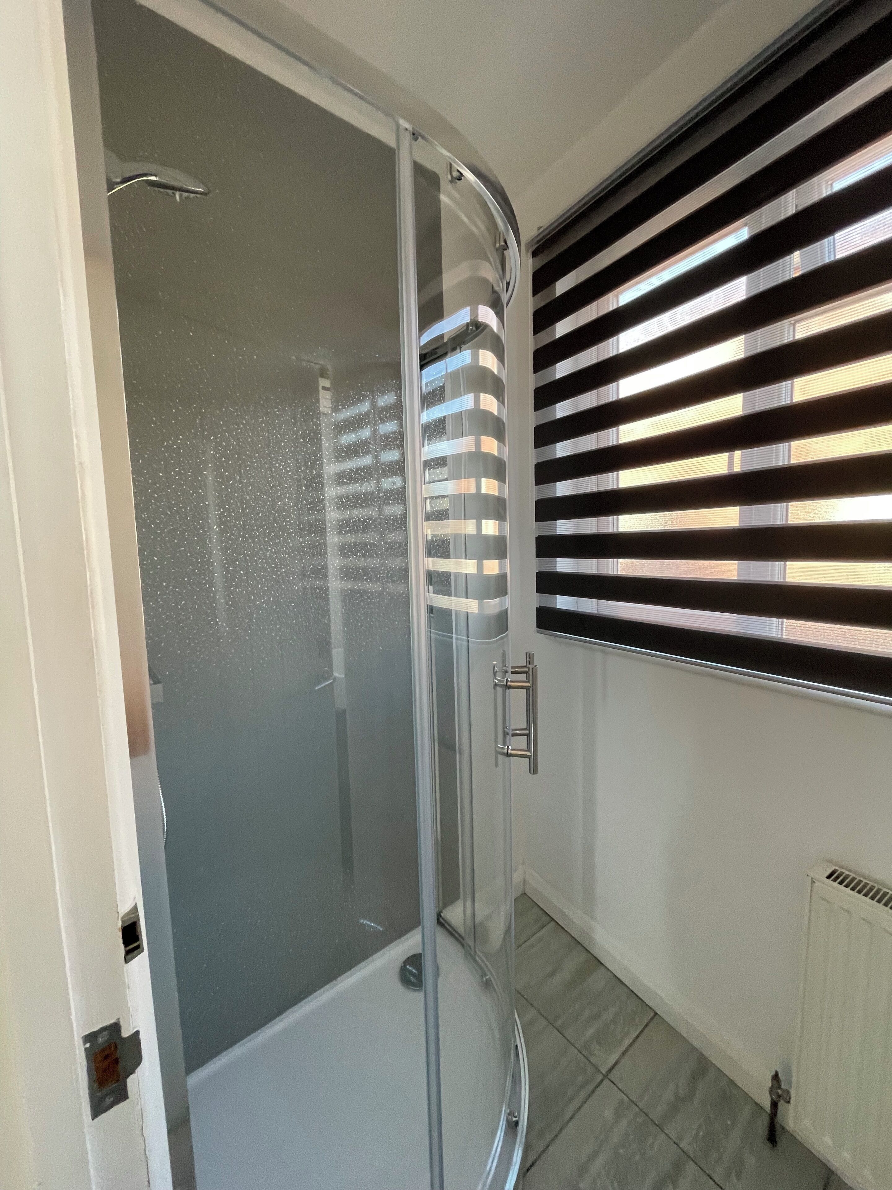 Un bagno moderno con cabina doccia in vetro, pavimento piastrellato di bianco e una finestra con persiane orizzontali.
