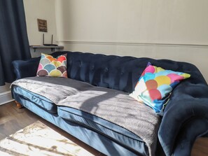 Ein dunkelblaues Sofa mit grauer Decke und bunten Kissen, ein kleiner Tisch mit einem Schild und ein Fenster mit Vorhängen.