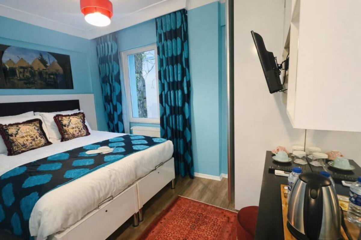 A bedroom with a bed, a window with curtains, a desk with a kettle, and a painting on the wall.