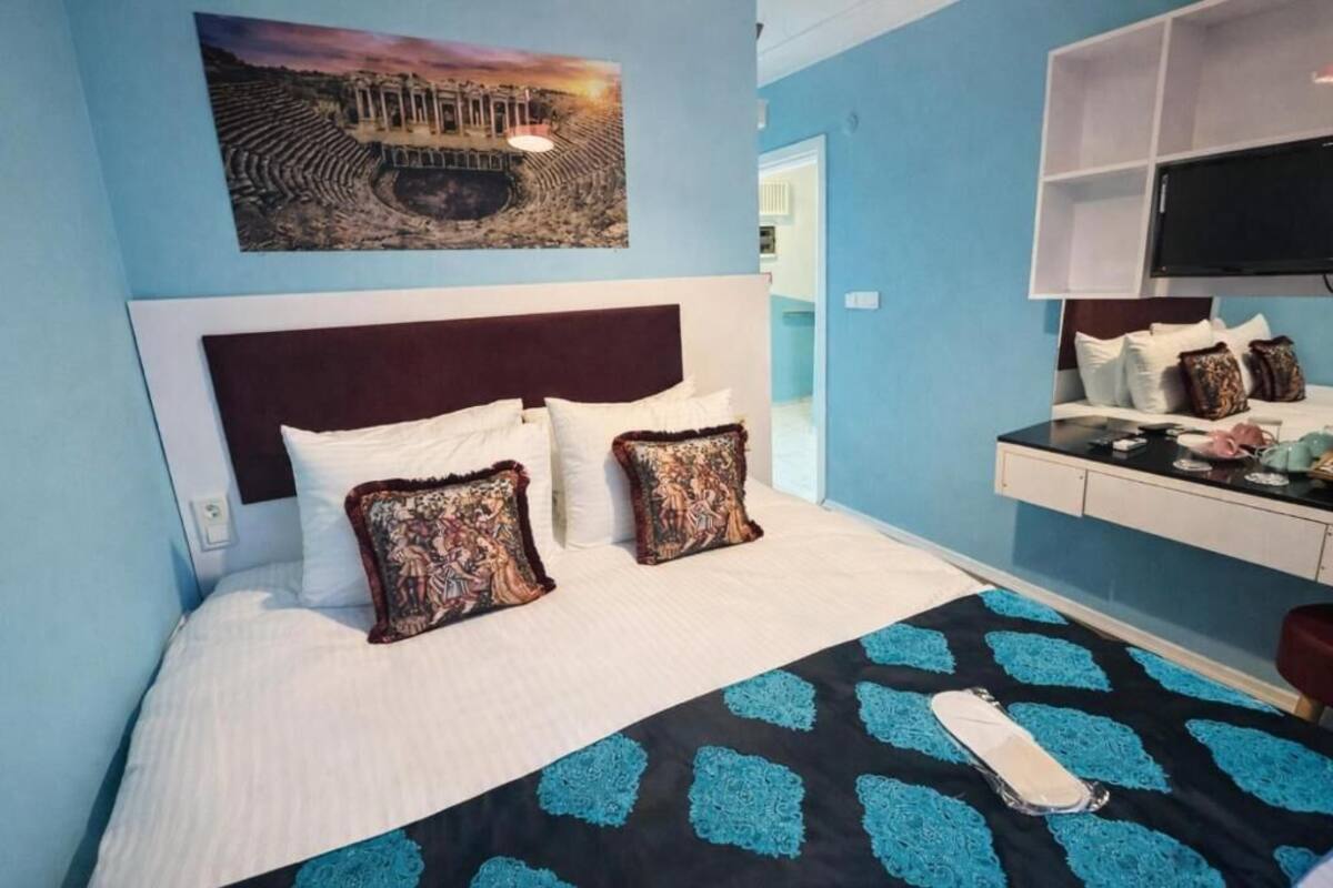 A hotel room with a bed, a flat-screen TV, a painting of an ancient amphitheater, and a blue and black patterned bedspread.