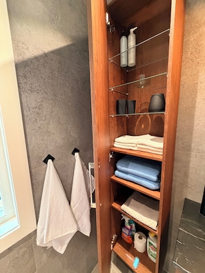 A bathroom with a wooden shelf holding towels, toiletries, and cleaning supplies. - A cozy vacation home surrounded by nature, with top-notch amenities! (Sotebyn)